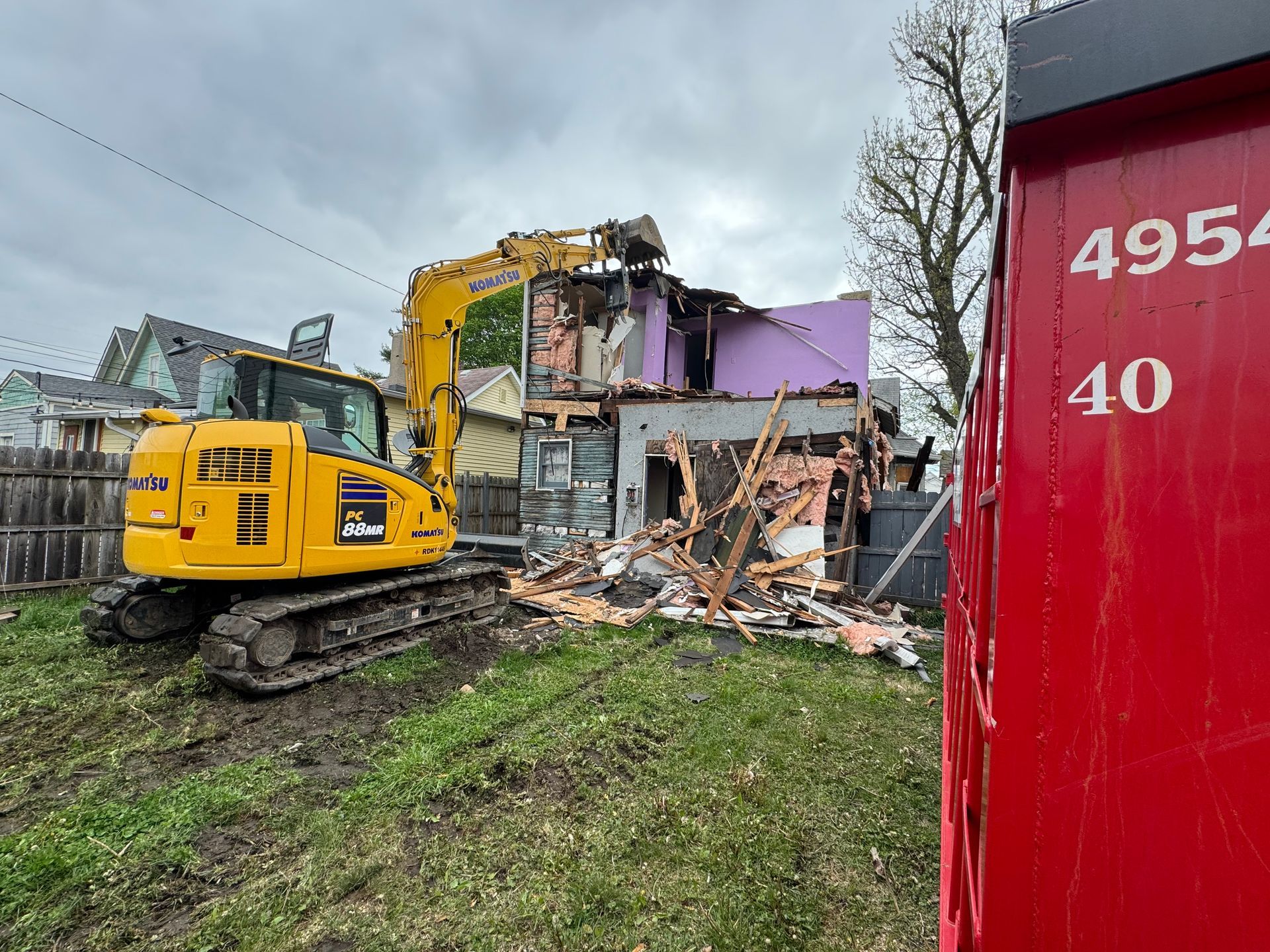 A yellow excavator is demolishing a house next to a red container.