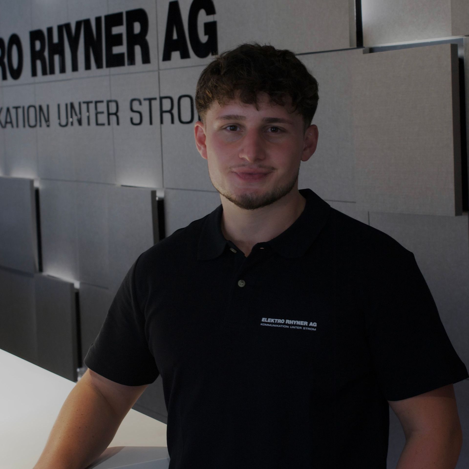 Ein Mann in einem schwarzen Hemd steht vor einem Schild mit der Aufschrift „Elektro Rhyner AG“