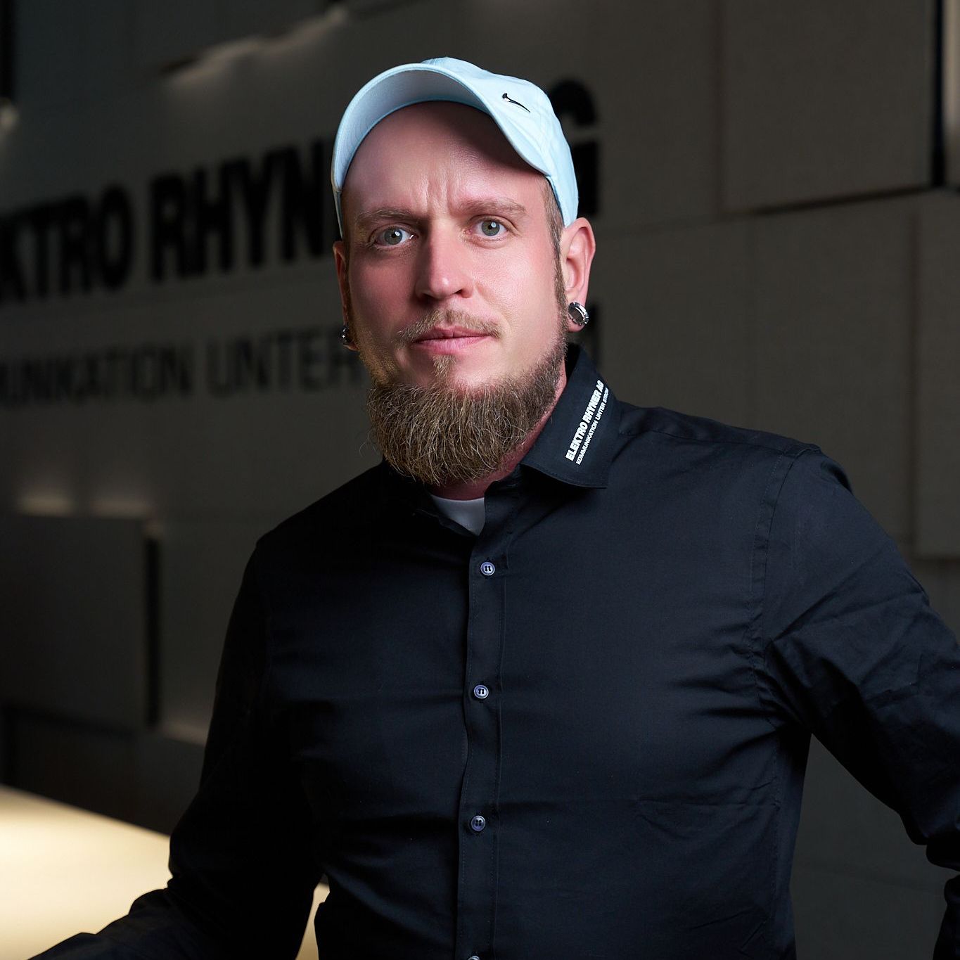 Ein Mann mit Bart trägt ein schwarzes Hemd und steht vor einer Wand mit der Aufschrift „Elektro Rhyner AG“