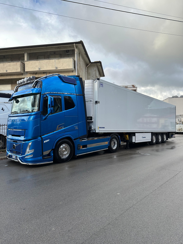 Un camion blu con rimorchio bianco percorre una strada bagnata accanto a un edificio industriale.