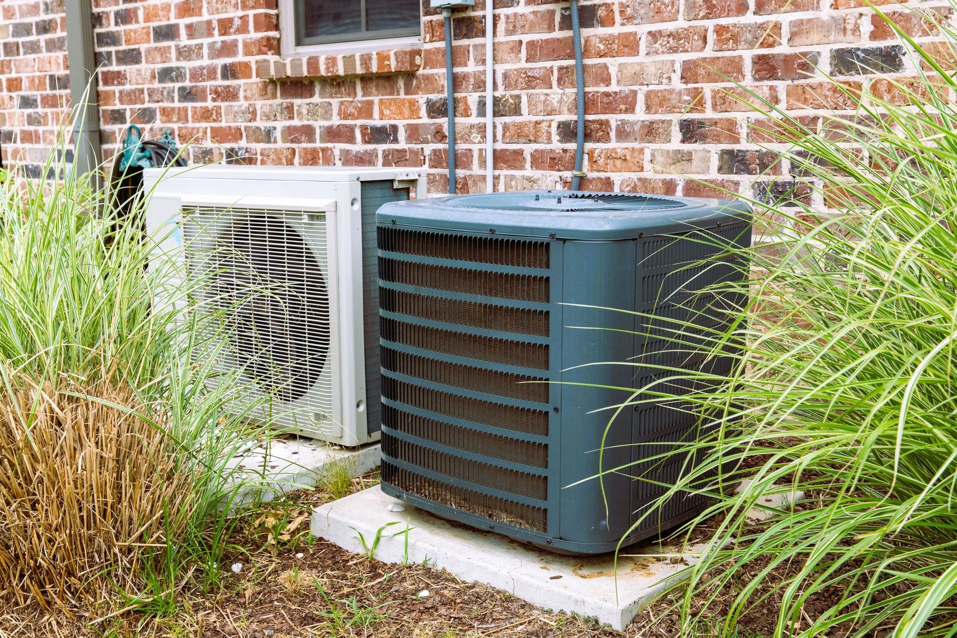 HVAC units outside a brick house surrounded by overgrown grass