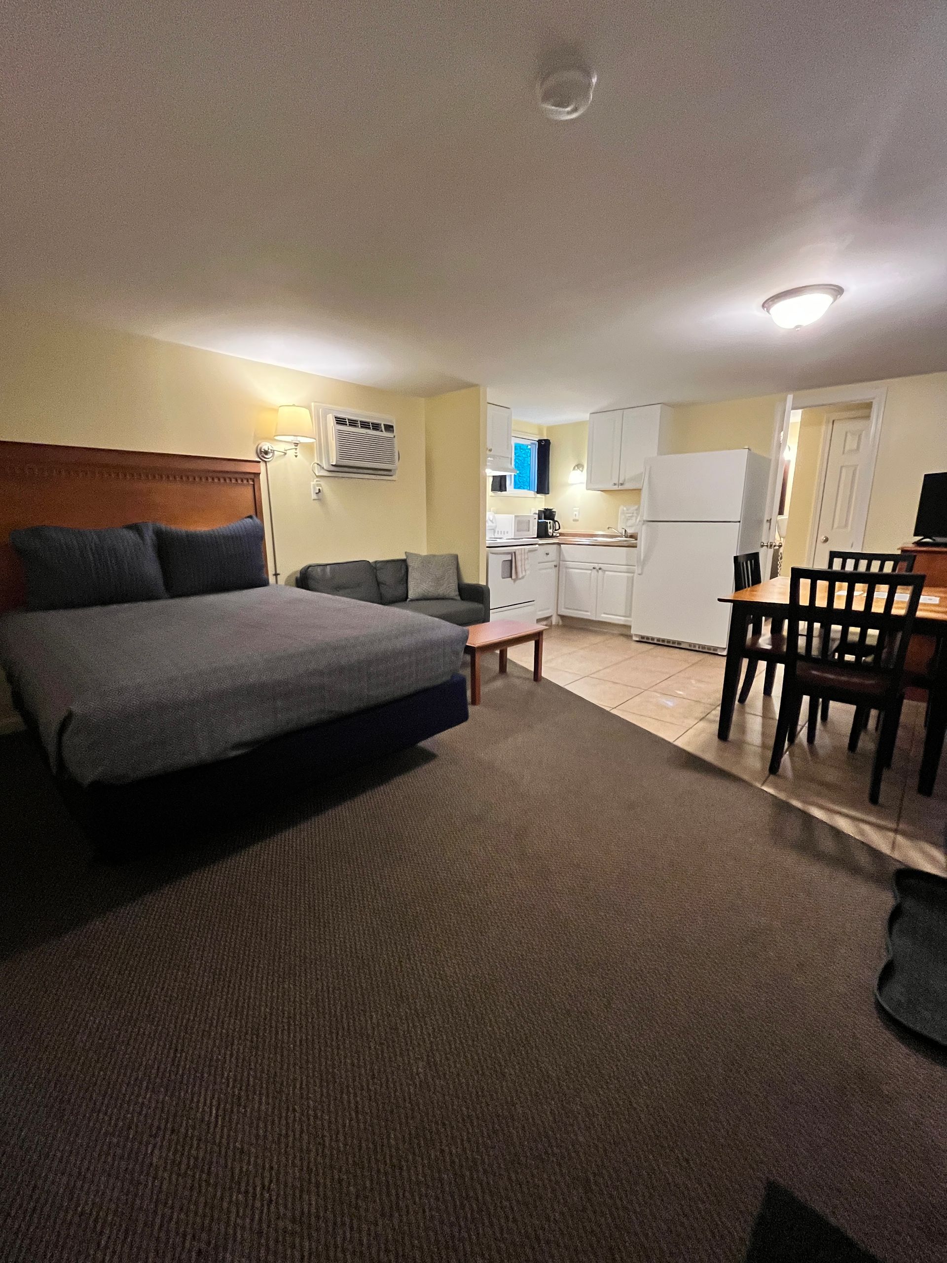 Bedroom with king bed, sofa, kitchen, and dining area. Neutral colors with brown carpet.