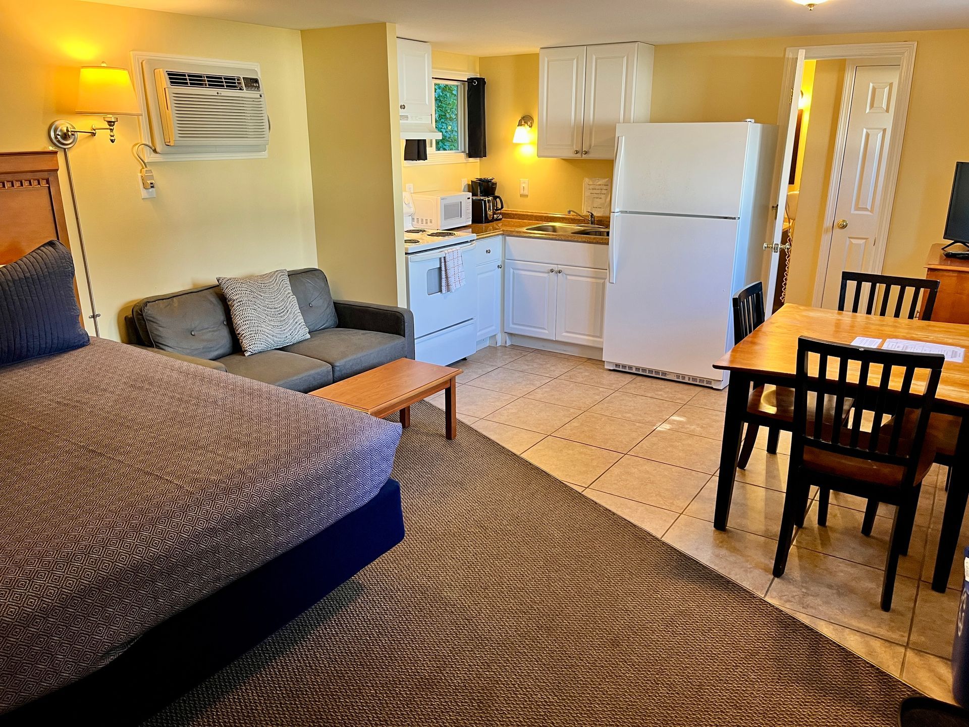 Studio apartment: bed, sofa, small kitchen with white appliances, dining table with chairs.