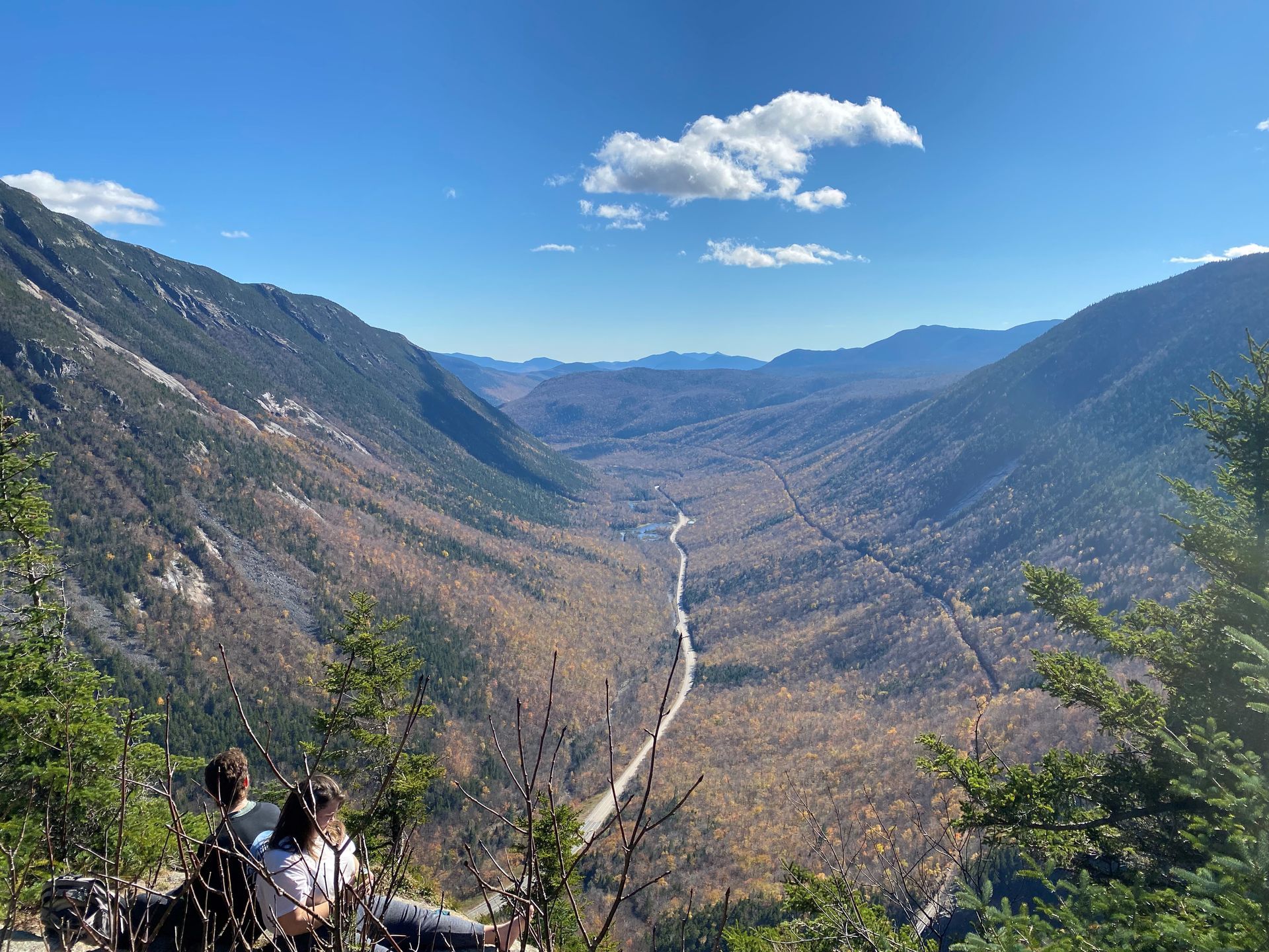 A couple of people are sitting on top of a mountain overlooking a valley.