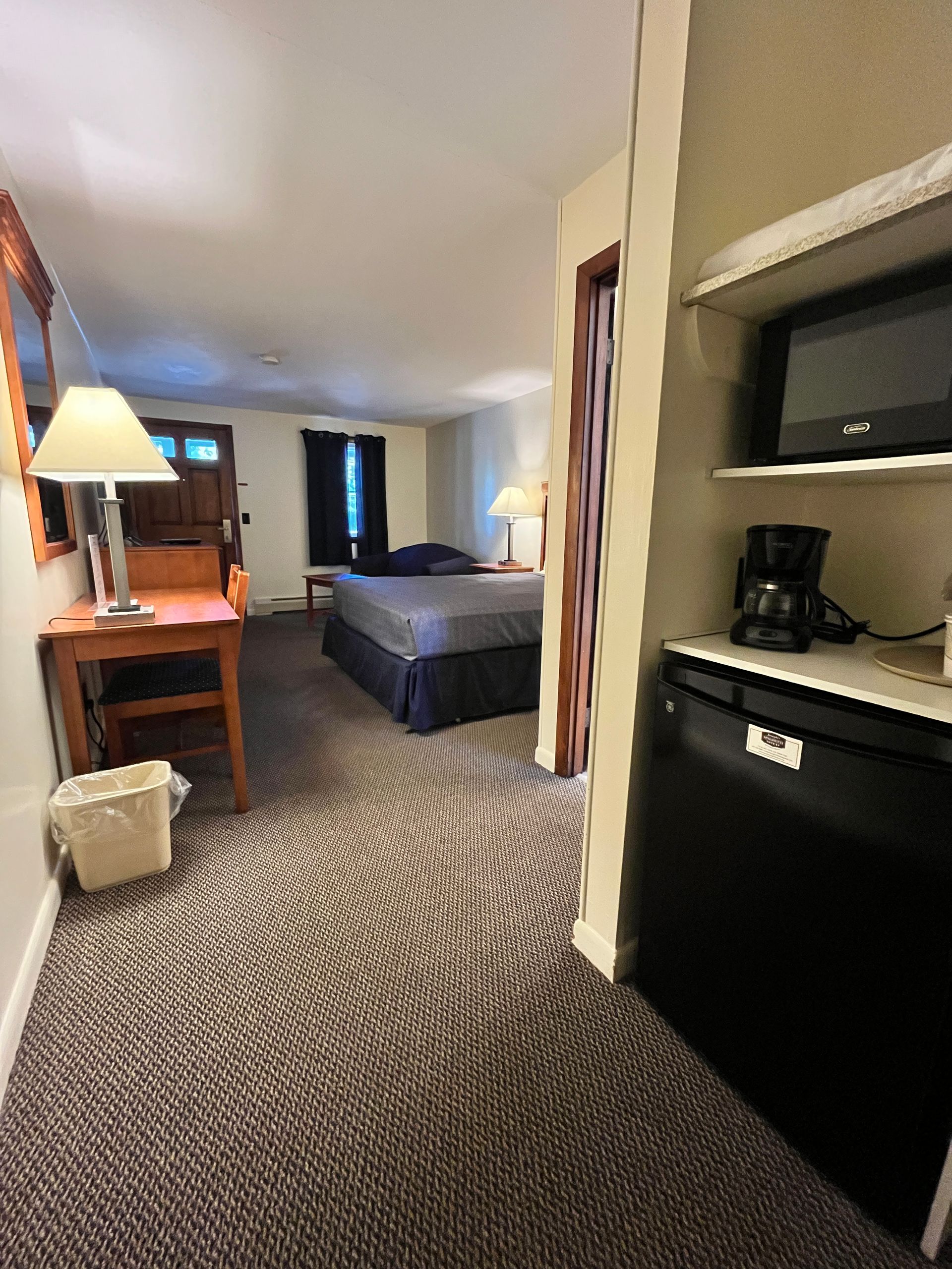 Hotel room interior with bed, desk, and microwave on shelf above mini-fridge. Carpeted floor.