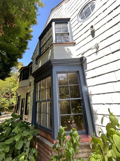 White clapboard house with black-trimmed bay windows and a brick foundation beside leafy greenery