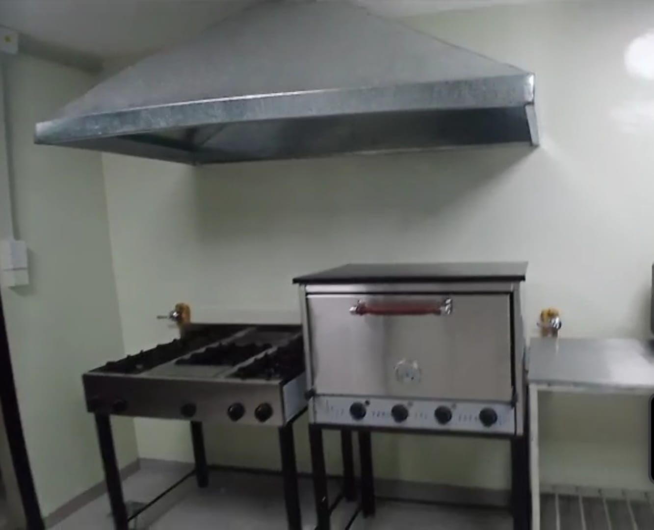 Cocina industrial con estufa, horno y campana extractora de gran tamaño de acero inoxidable en una pared blanca.
