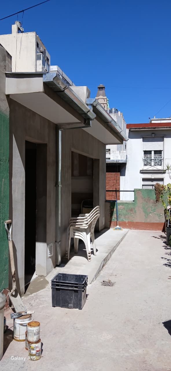 Un estrecho patio soleado junto a un edificio, con sillas, cubos y una mesita a lo largo de un sendero de cemento.