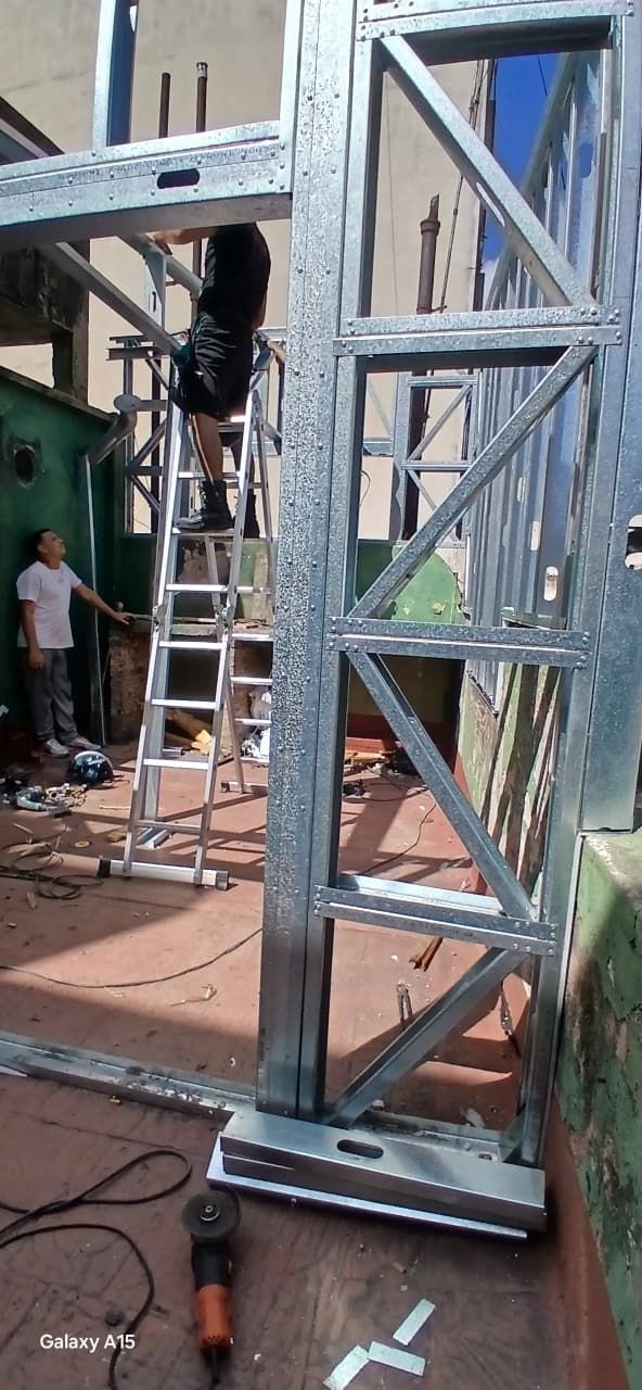 Un trabajador subido a una escalera ensambla una gran estructura metálica gris en una obra de construcción.