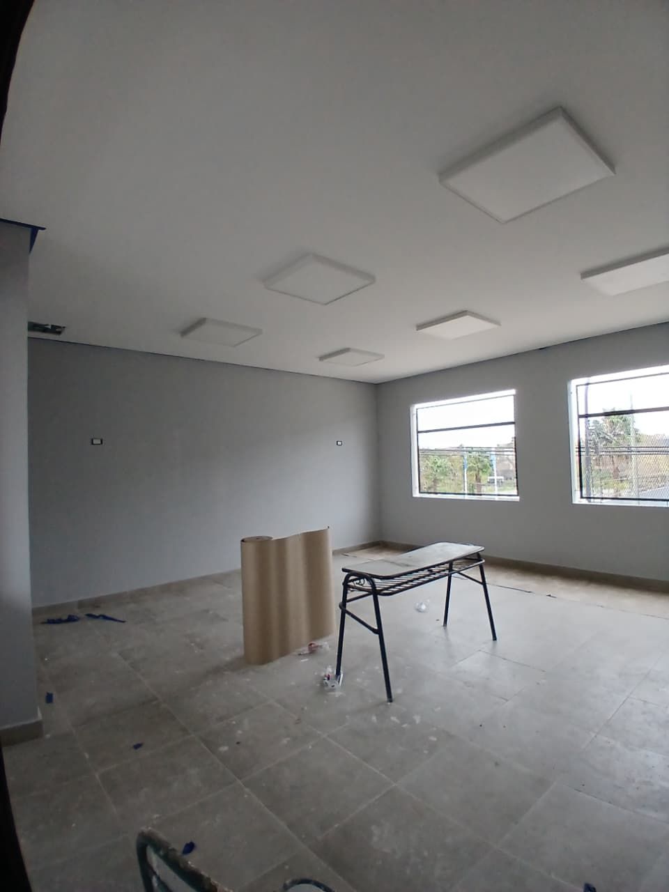 Habitación vacía con paredes grises, suelo de baldosas, ventanas y una mesa de trabajo en el centro.