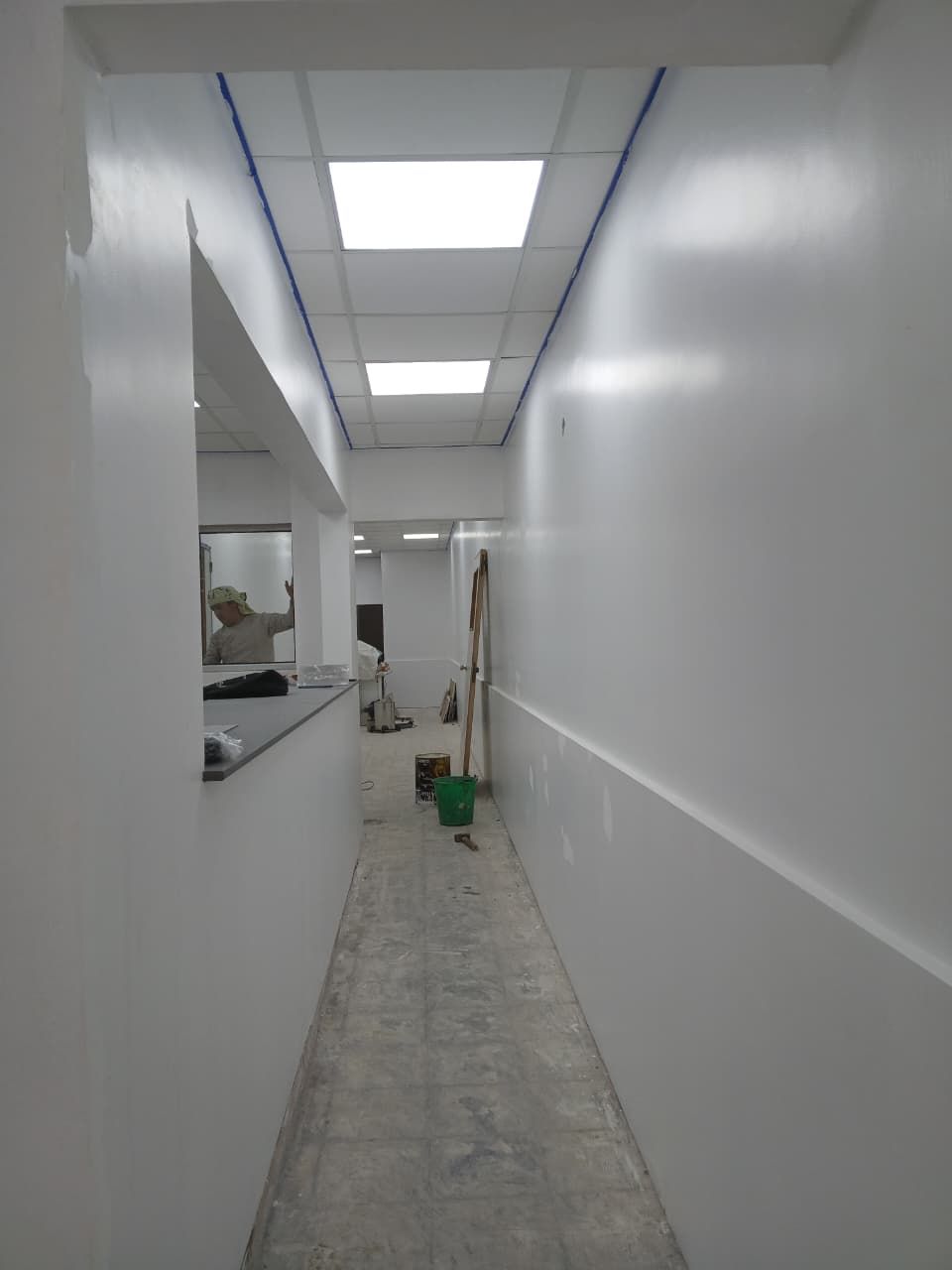 Un estrecho pasillo blanco con luces de techo, una ventana y productos de limpieza en el suelo.