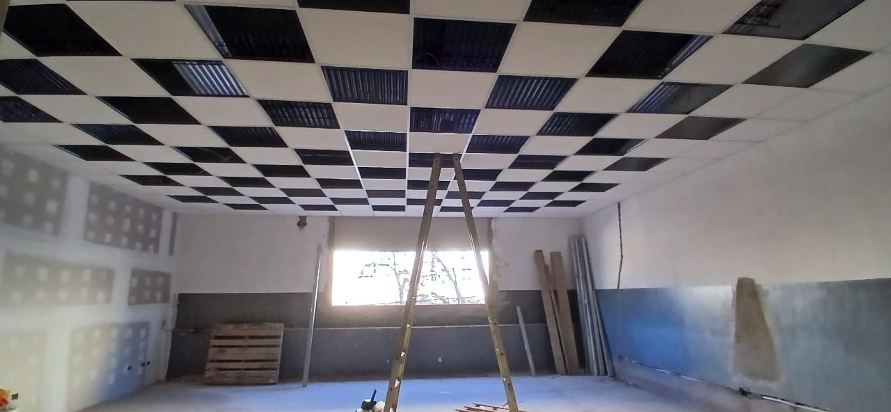 Habitación parcialmente pintada con techo a cuadros y escalera centrada bajo una ventana luminosa.