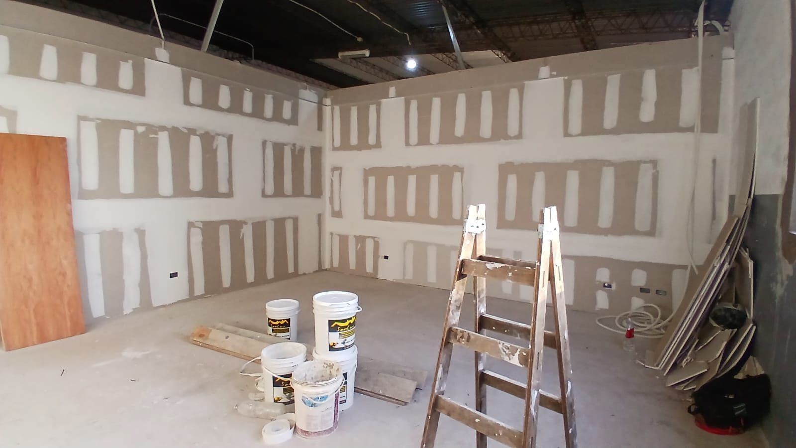 Habitación de obra con parches de paneles de yeso, cubos de pintura y una escalera sobre un piso de concreto.