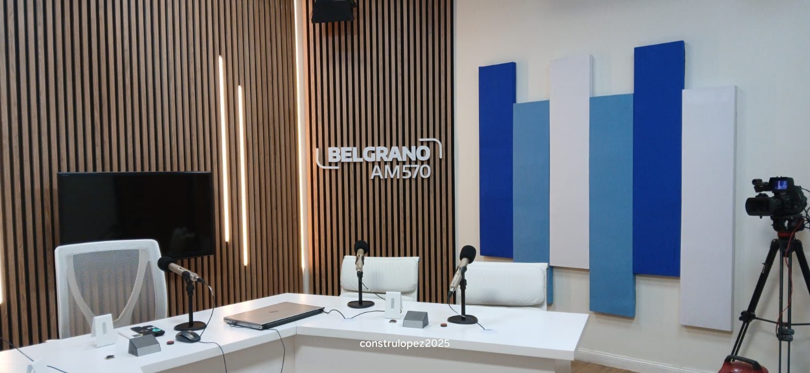 Estudio de podcast con micrófonos, mesa blanca, paneles acústicos azules y una cámara sobre un trípode.