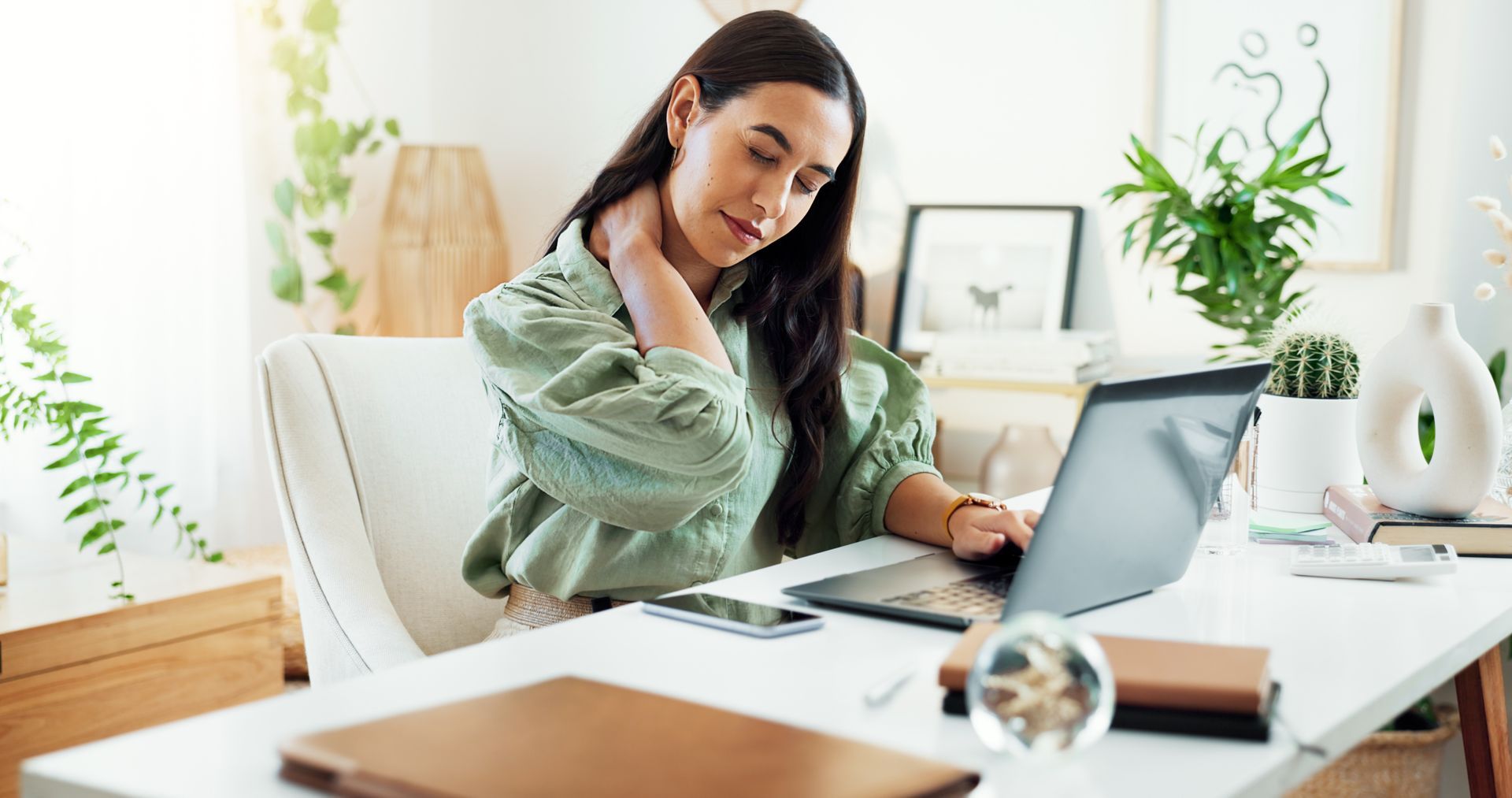 Woman working at a bright home office desk with a laptop, papers, and plants nearby