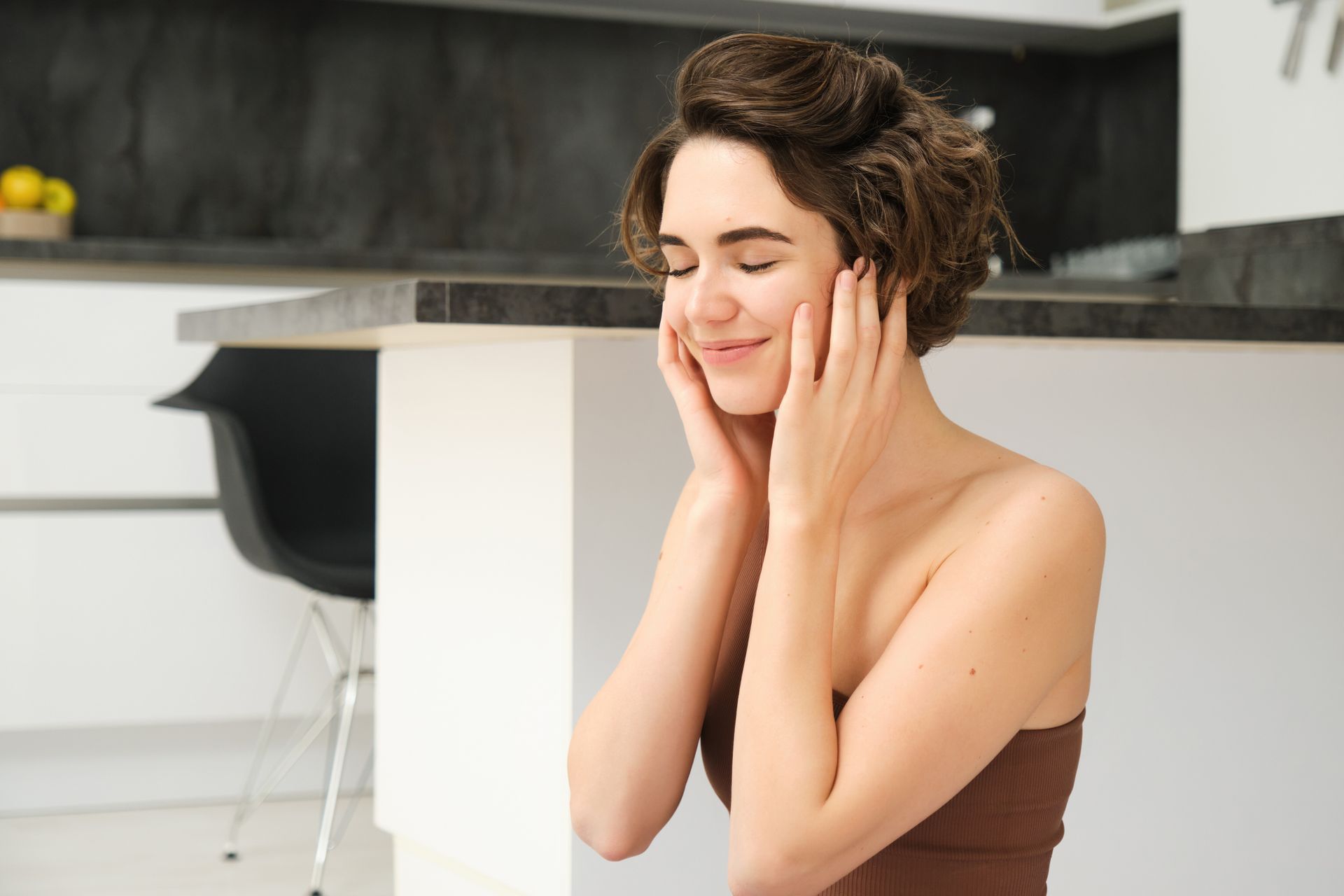 Woman with eyes closed, gently touching her face in a bright kitchen.