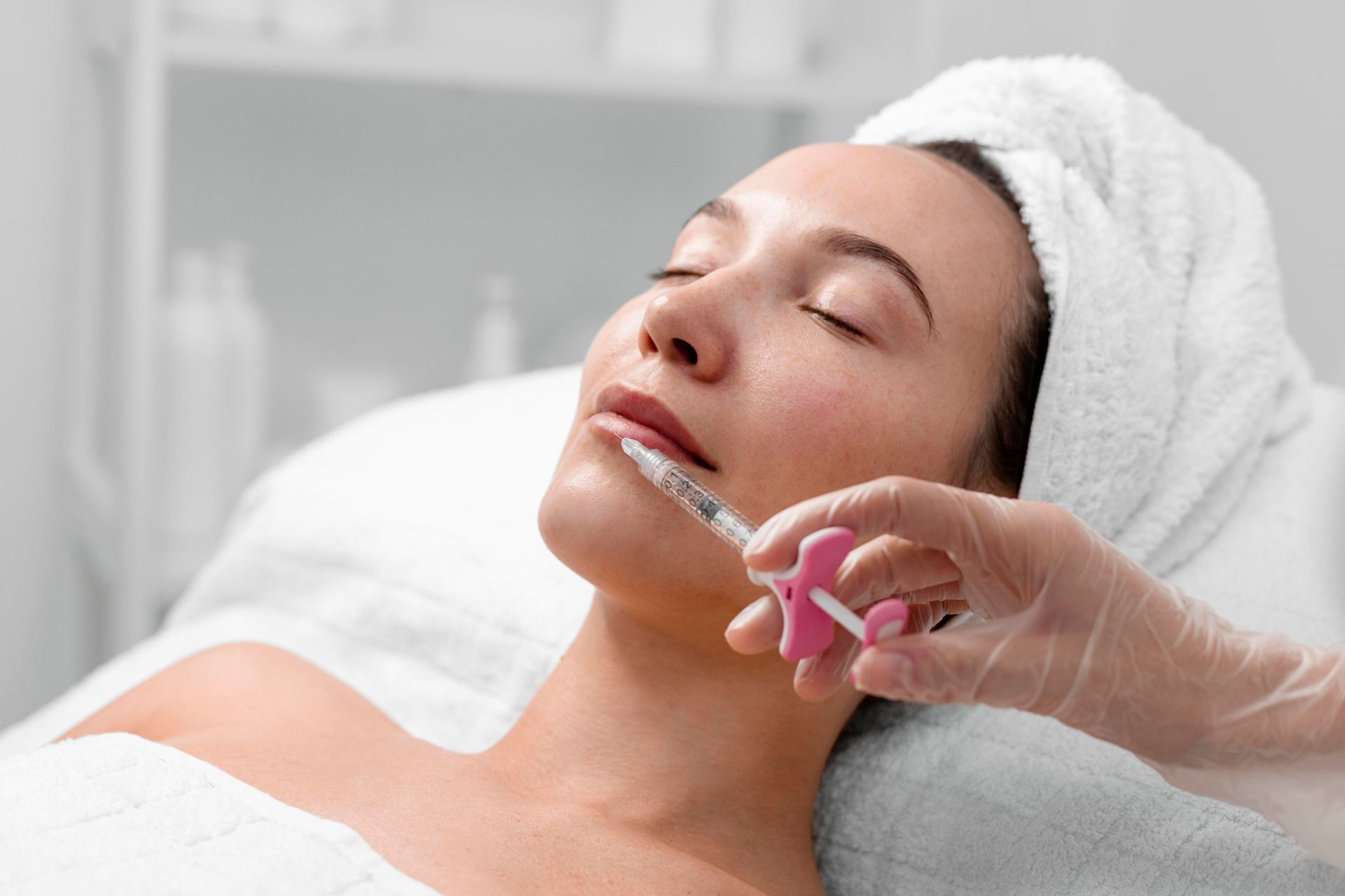 A gloved hand uses a syringe to inject cosmetic filler into a person's upper lip in a clinical setting.