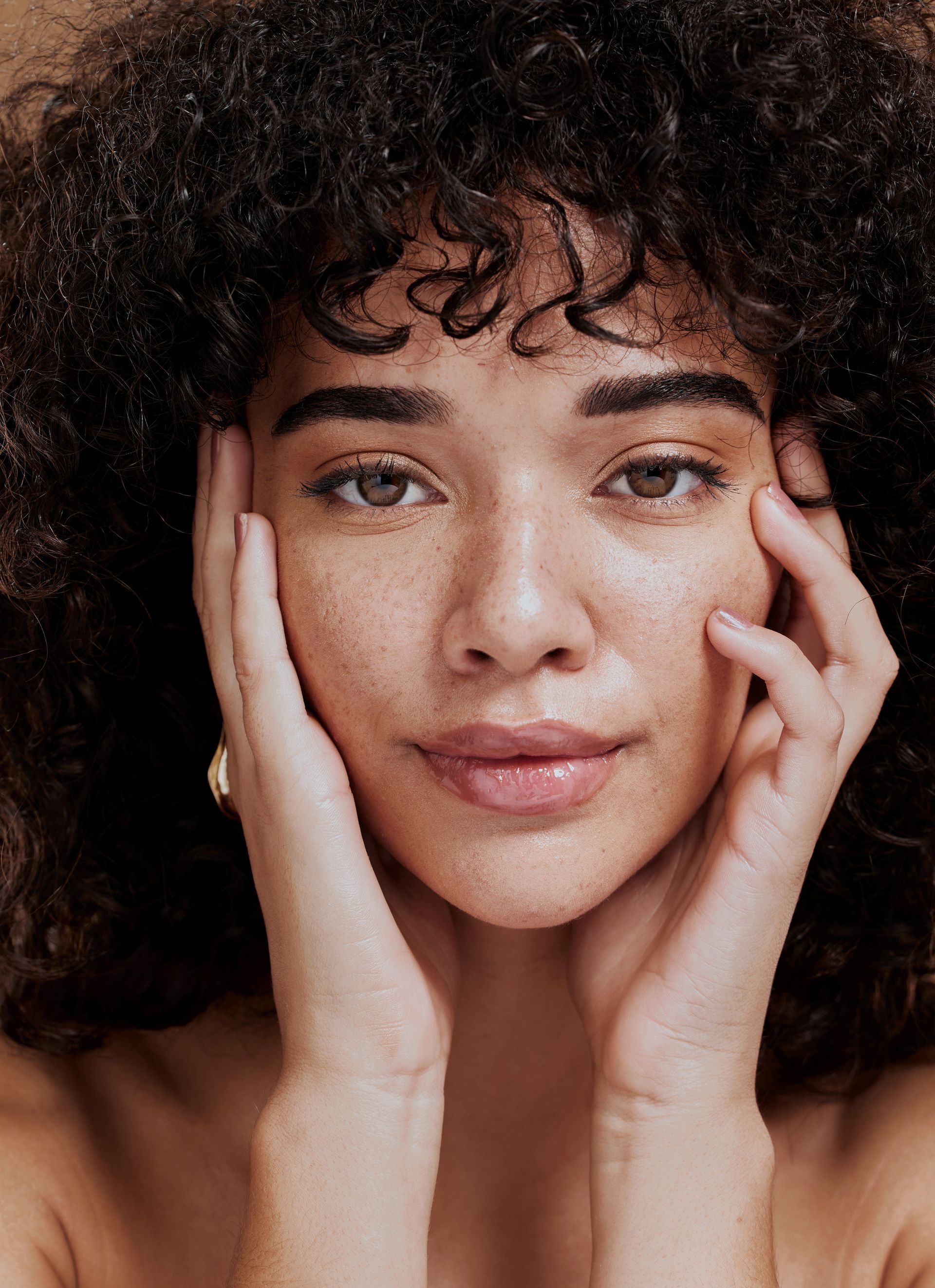 Close-up portrait of a person with curly hair, hands on cheeks, looking directly at the camera.