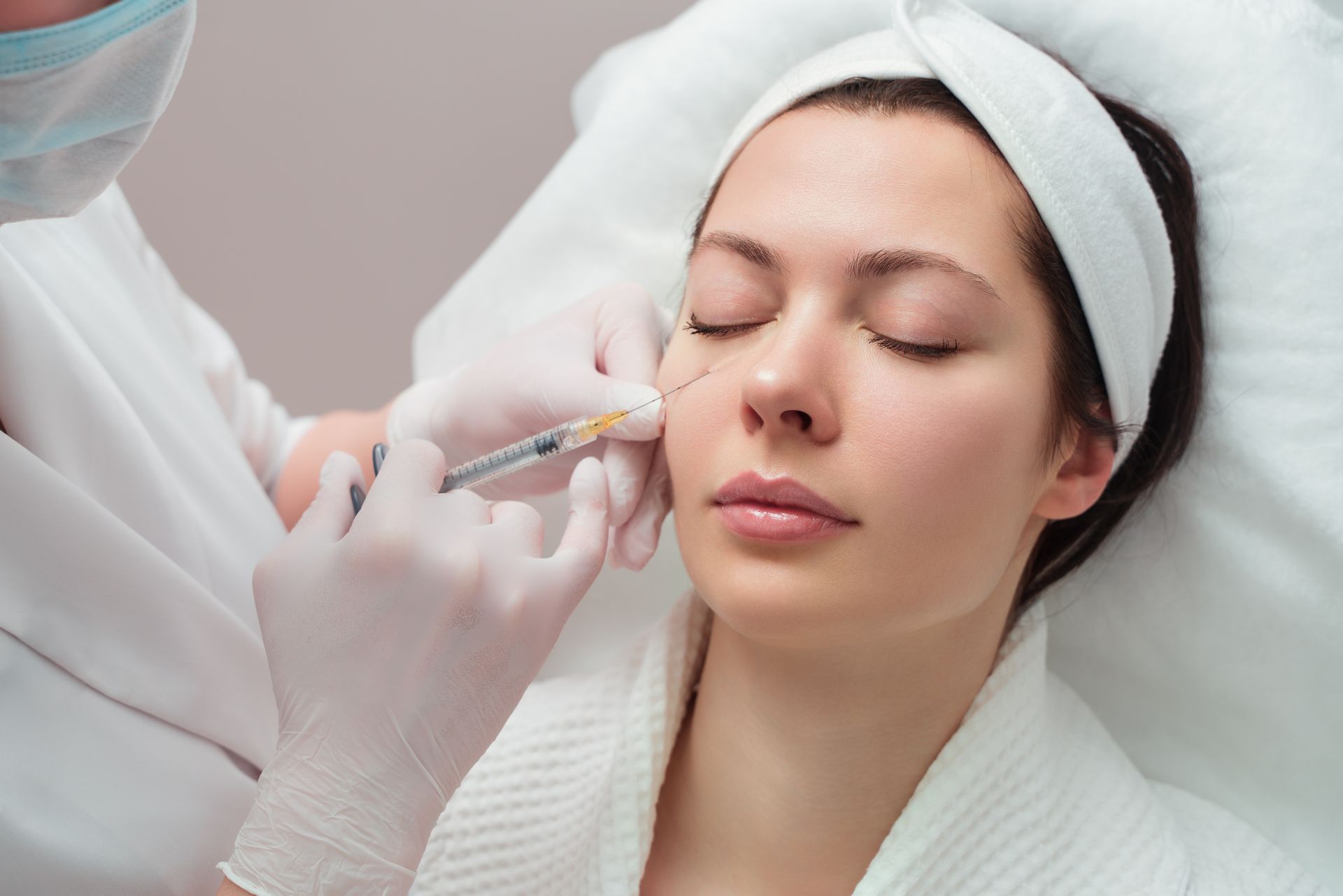 Cosmetic treatment to a woman’s cheek by a clinician using a syringe, with a white towel headband.