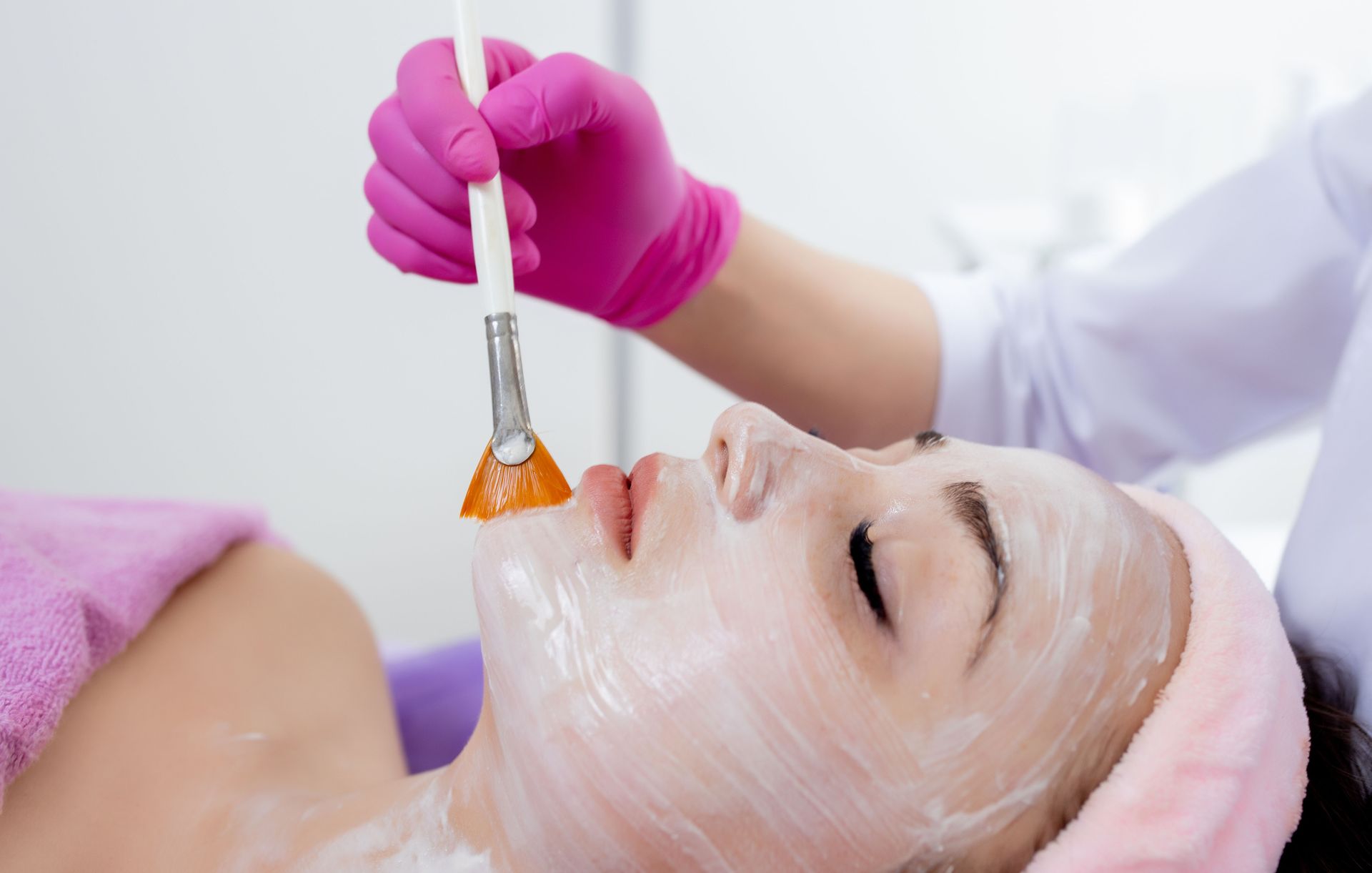 Gloved esthetician applying facial mask with a brush to a spa client lying on a treatment bed