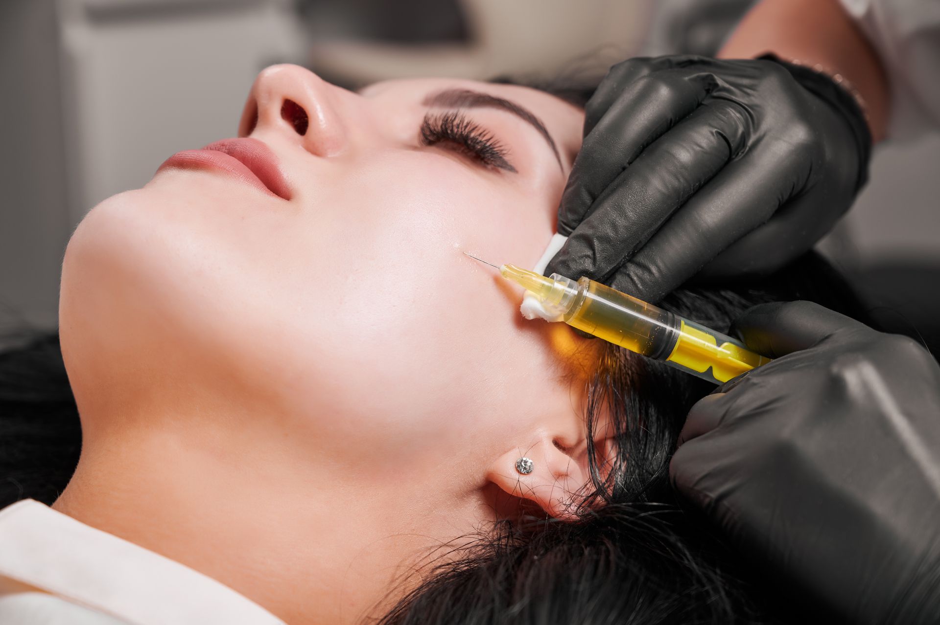 Person receiving a facial injection with a syringe while lying back, gloved hand holding skin aside