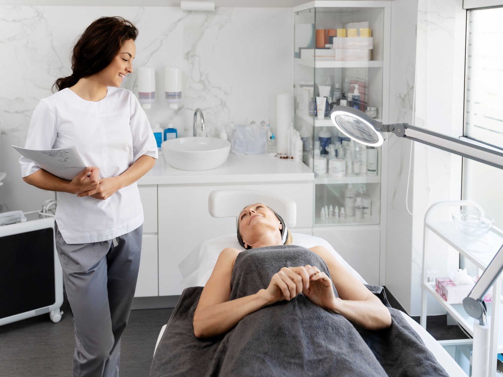 Esthetician consulting a client lying on a treatment bed in a bright spa room