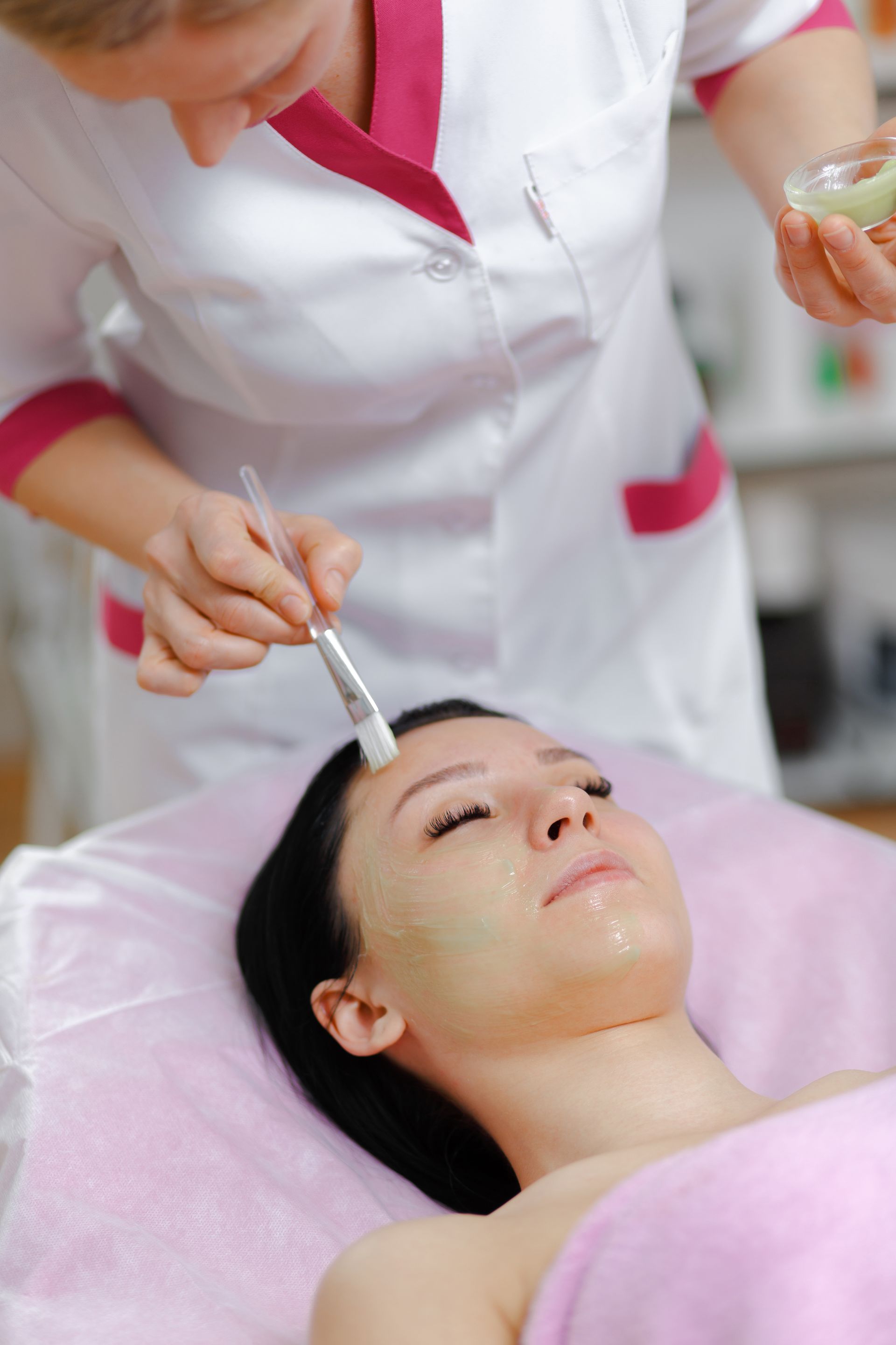 Esthetician applying facial treatment to a woman lying on a pink spa bed