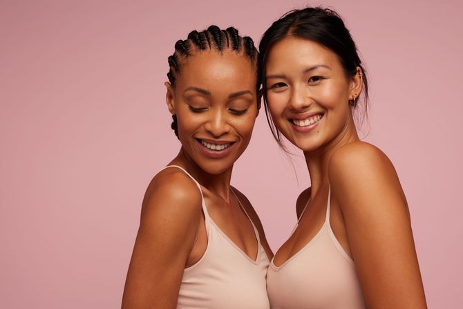 Two smiling women in beige tops posing against a pink background