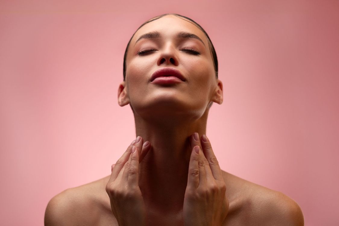 A person with glowing skin and a low ponytail resting their hand against their jawline against a plain gray background.