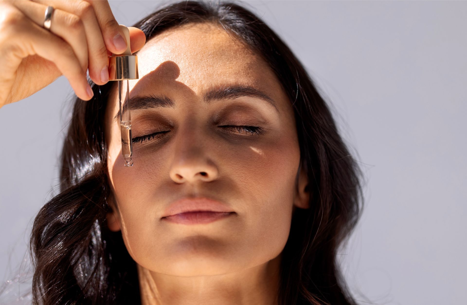 Woman applying facial serum dropper to her forehead, eyes closed, in a close-up skincare scene