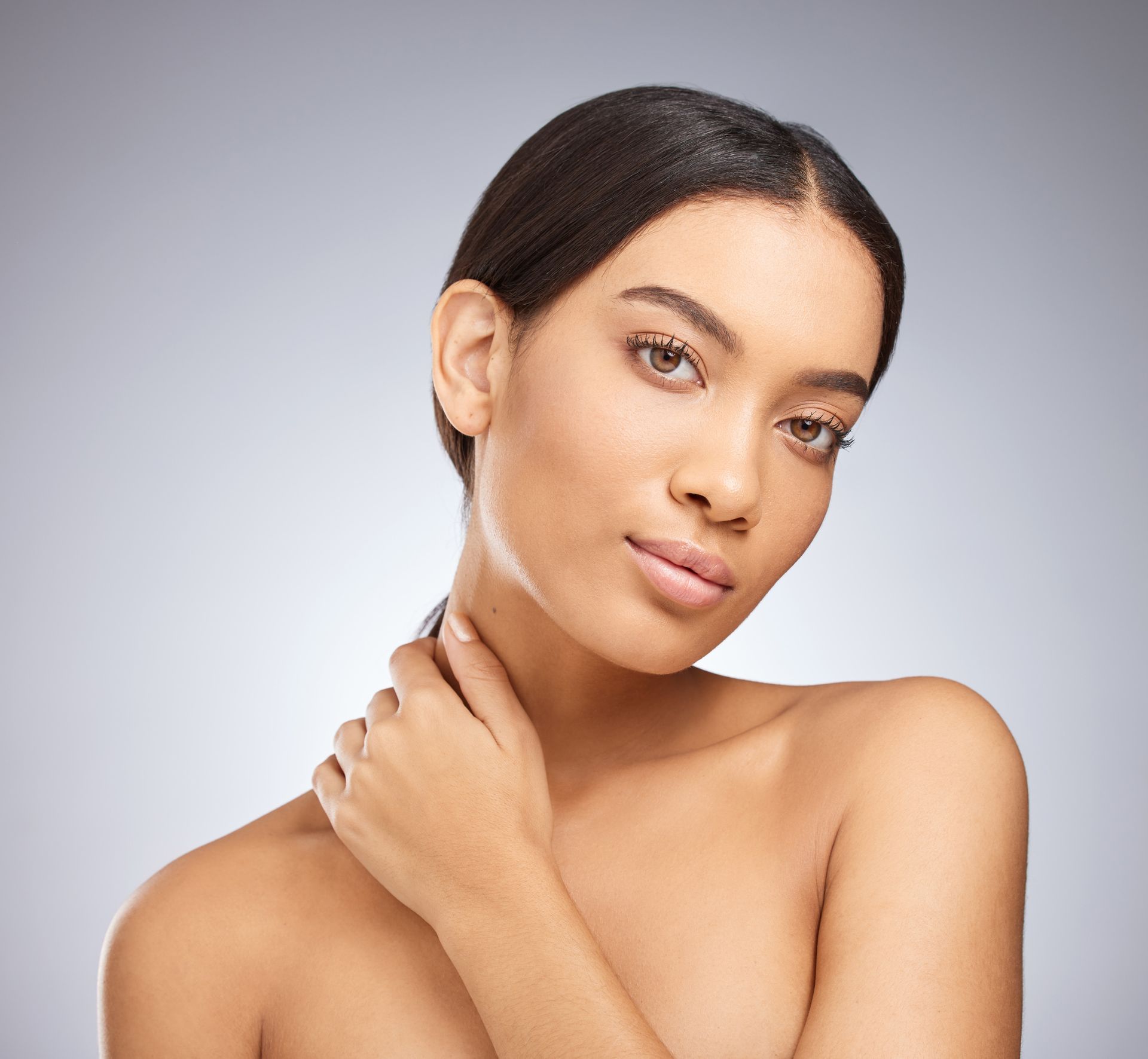 Portrait of a woman with dark hair against a light gray background, touching her neck and looking at the camera