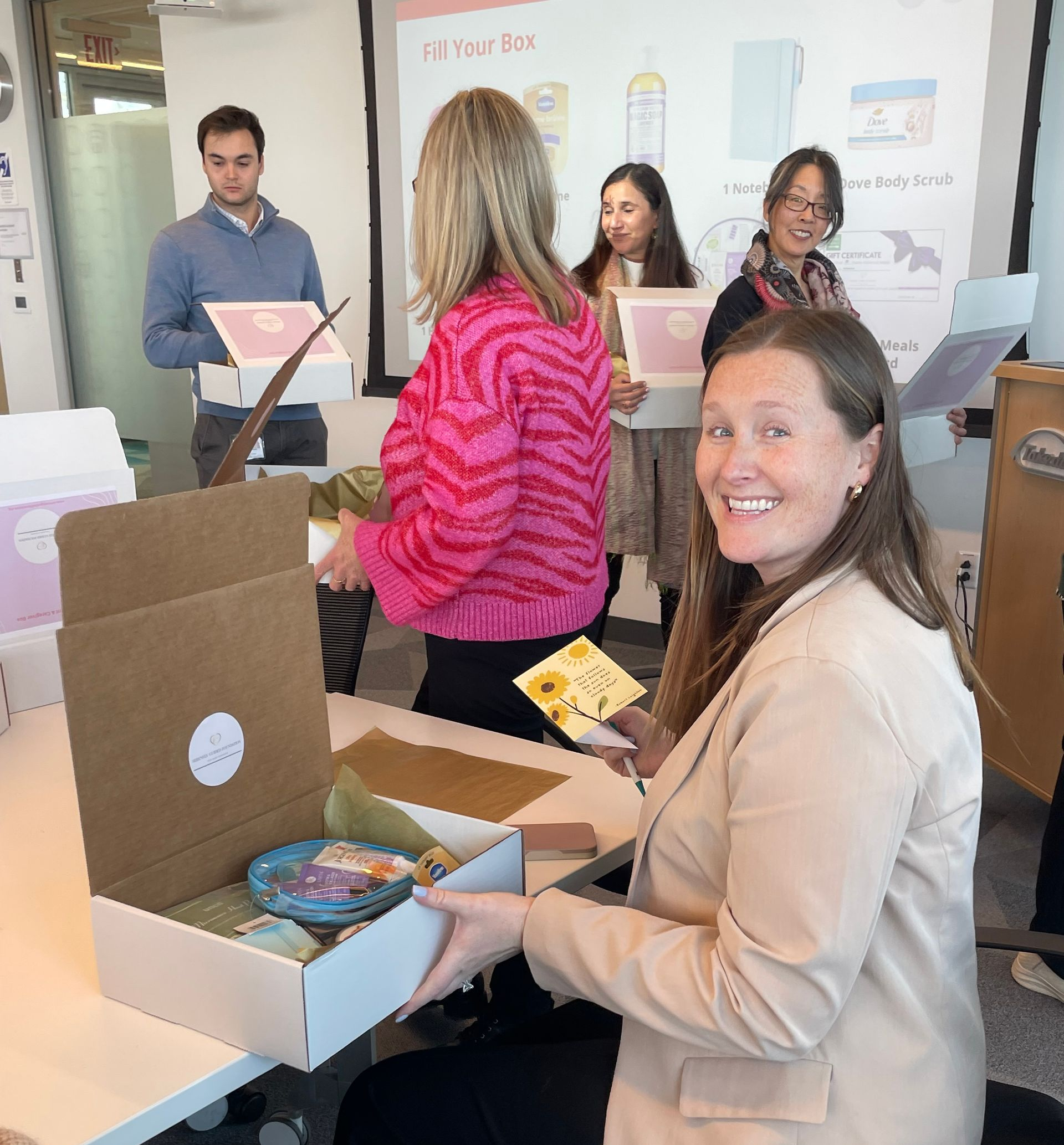 People gathered in a classroom opening gift boxes, with one smiling woman seated in the foreground.