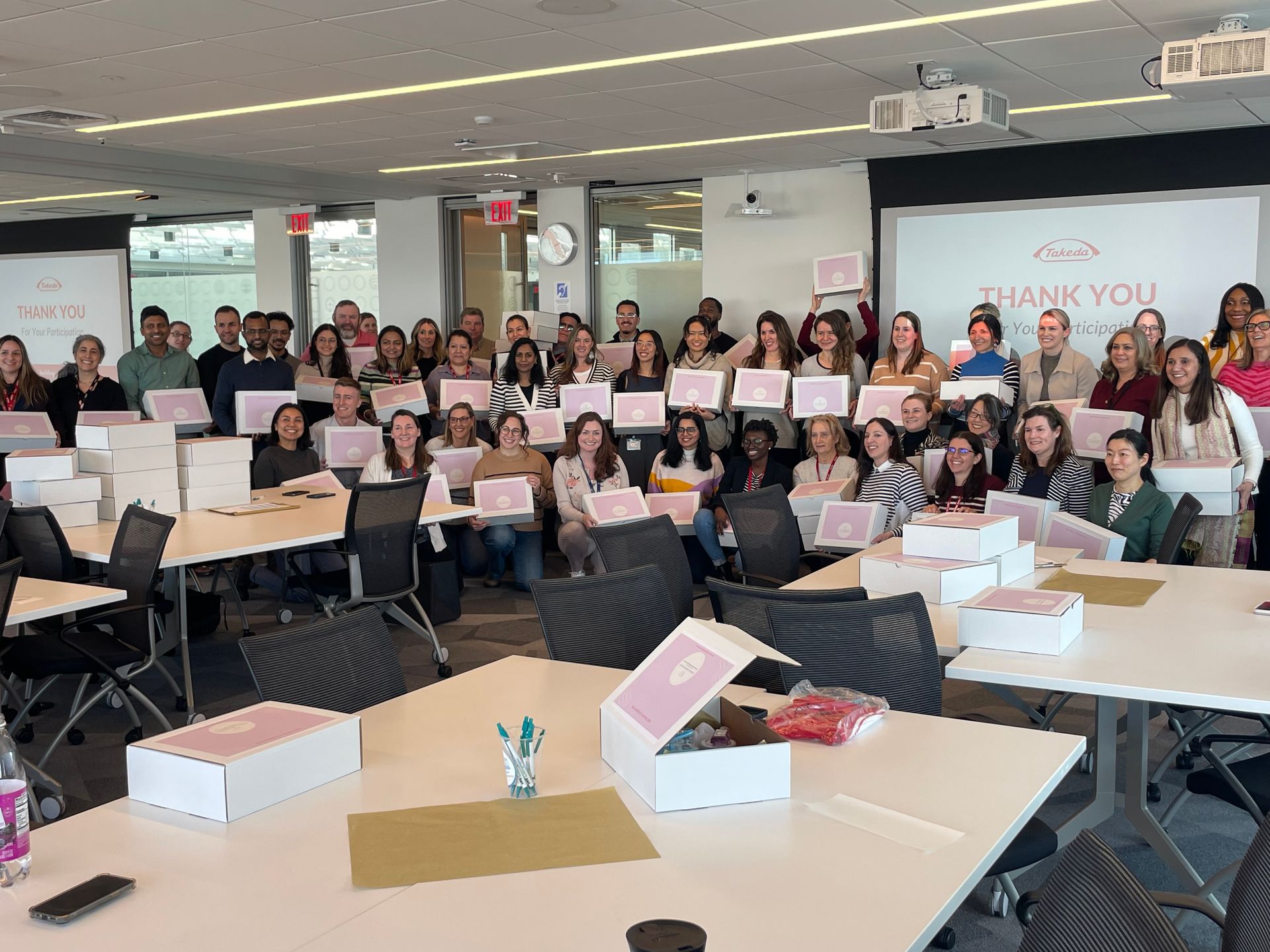 Large group posing indoors with white gift boxes and a “Thank you” banner in the background