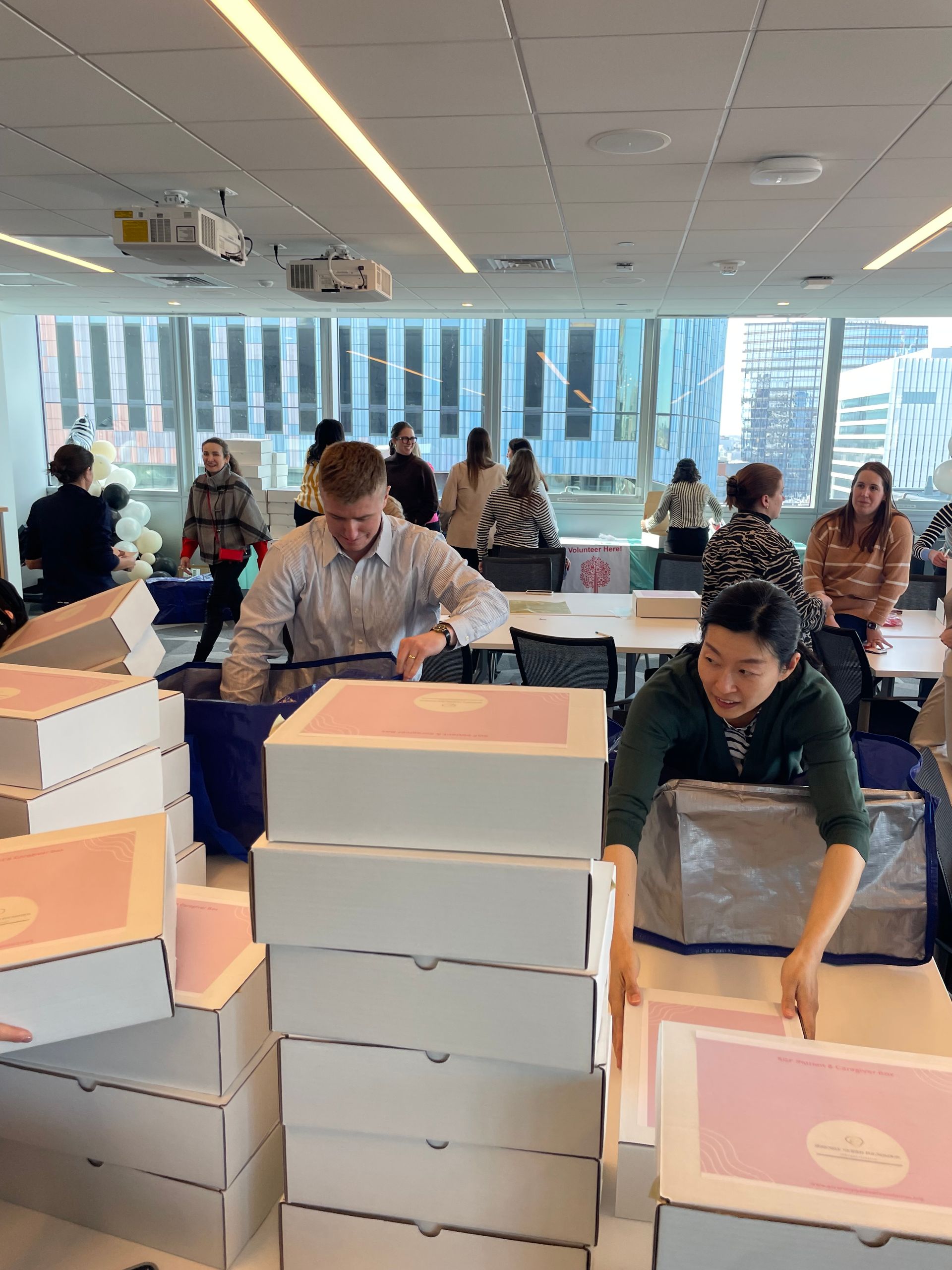 People stacking pink and white boxes in a bright office with city windows