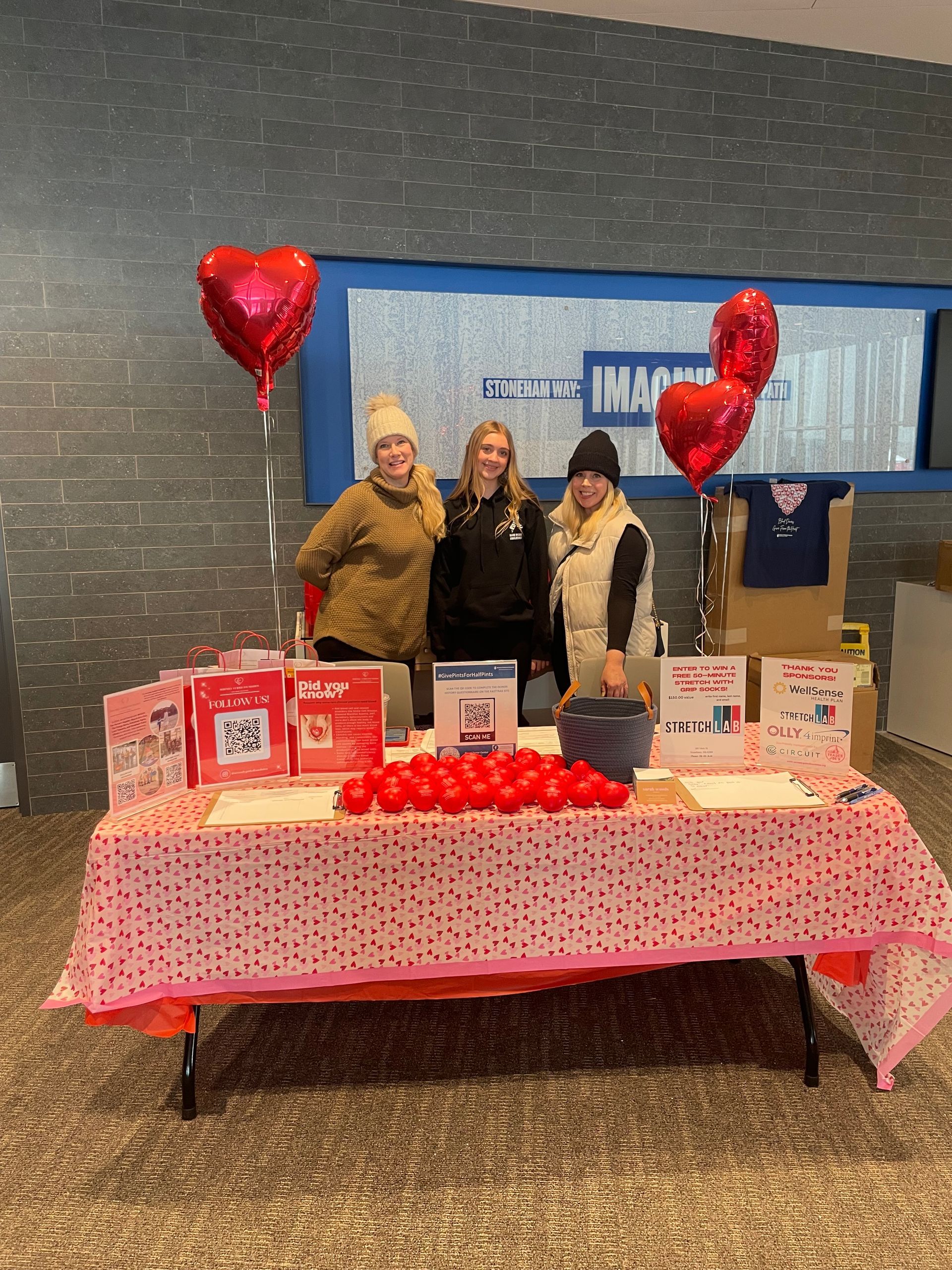 Three women are standing in front of a table with balloons and roses on it.