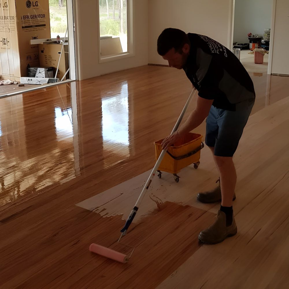 A Man Is Painting A Wooden Floor With A Roller And A Box — Kyall Chalker Floor Sanding & Polishing In North Haven, NSW
