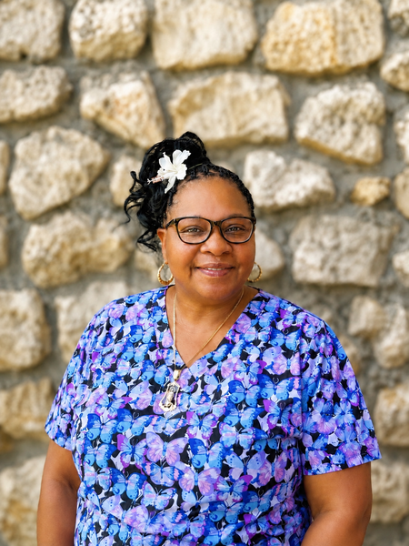 Woman in a blue floral blouse and glasses smiling before a stone wall.