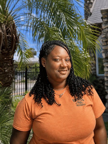 Person in an orange shirt smiling outdoors near palm trees and a stone building.