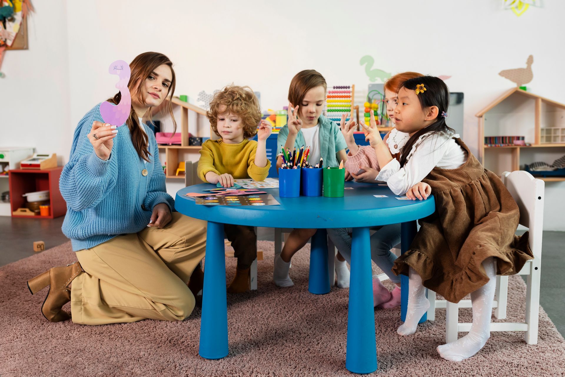 Children painting together at a blue table in a cozy classroom, with colorful art supplies and craft paper.