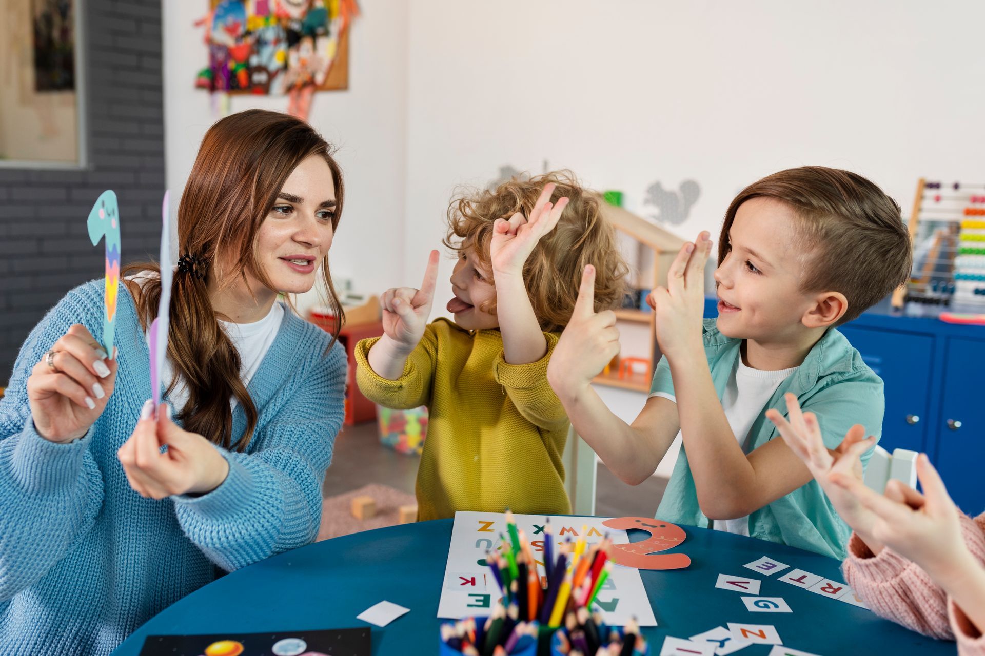 Children crafting with colorful supplies at a table, smiling and raising their hands in a bright classroom.