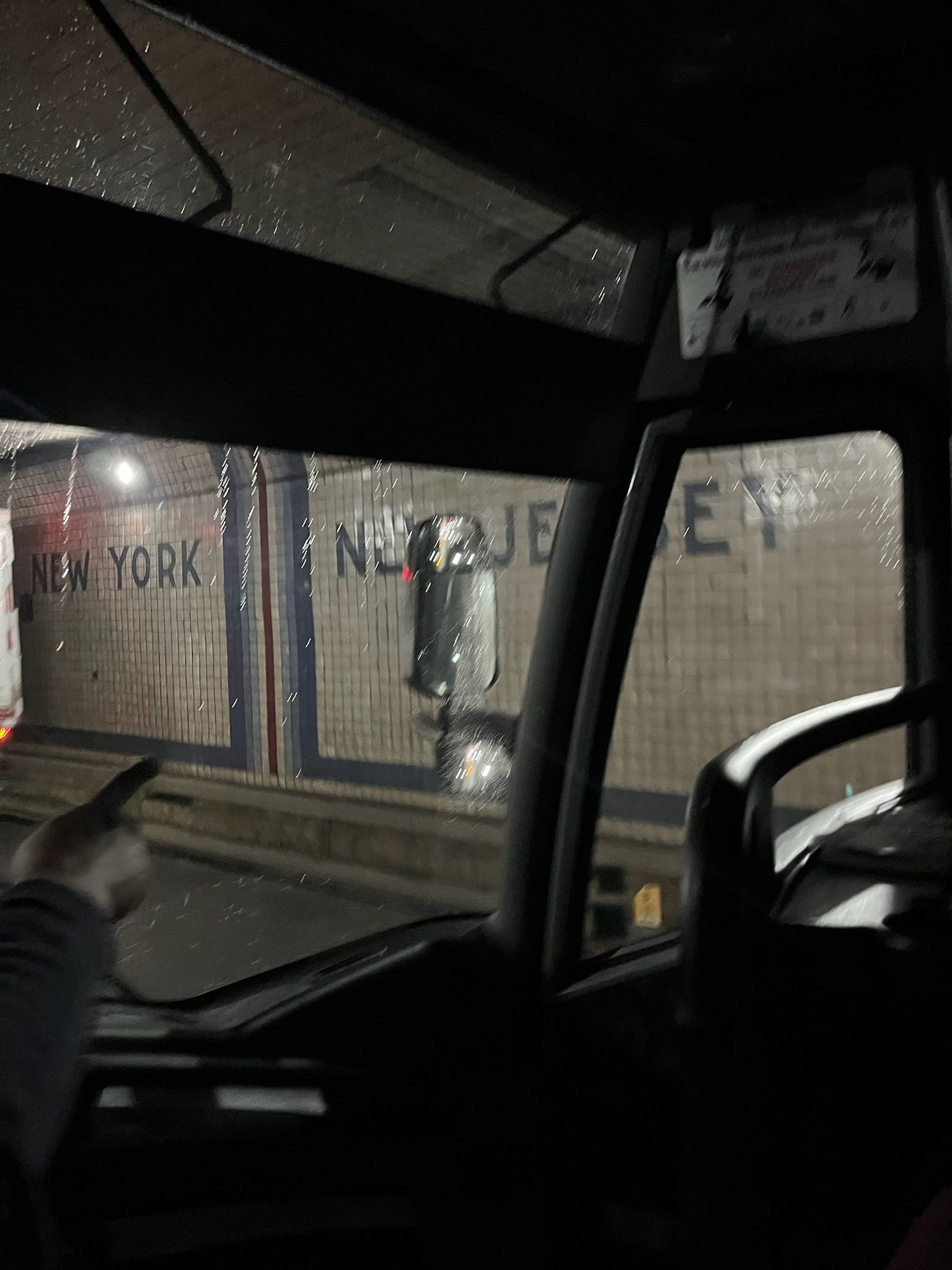 Rainy street view through a vehicle windshield at night, with a lit storefront and parked cars outside.