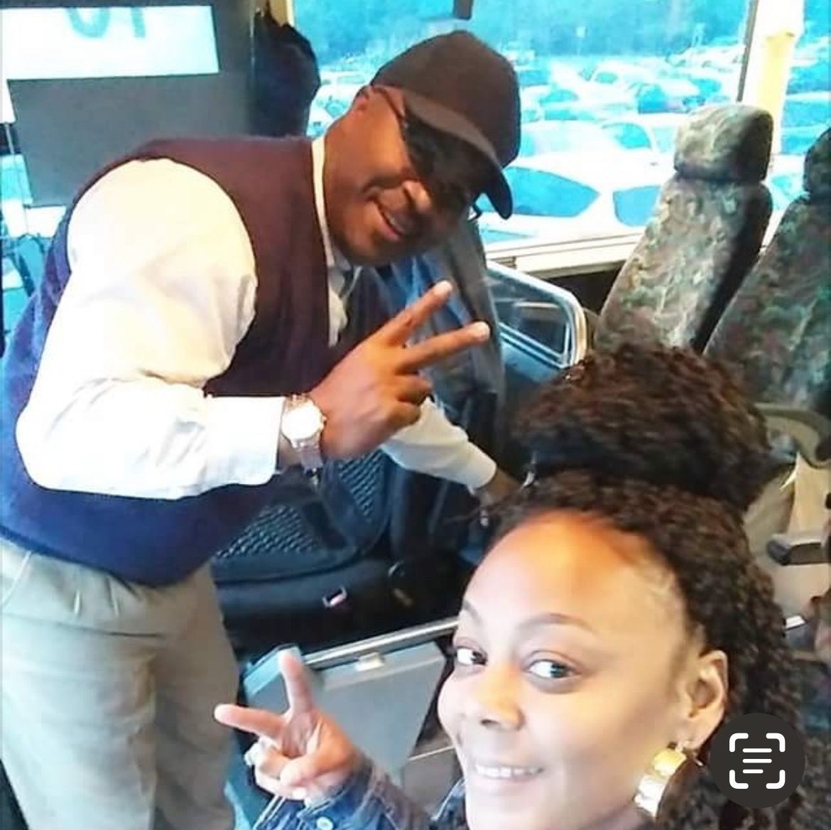Two people smiling and flashing peace signs on a bus, with seats and a window in the background.