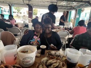 Two people smiling at a restaurant table with seafood and drinks, other diners visible in the background.