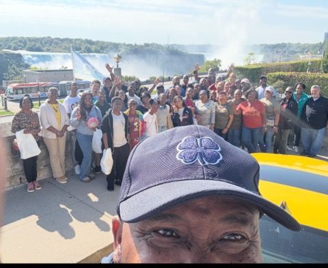 Crowd gathered on a roadside under blue sky, with a man’s cap and face in the foreground.