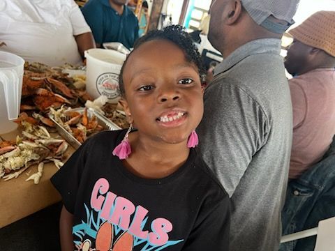Smiling child in a black T-shirt stands by a table with food and people seated nearby