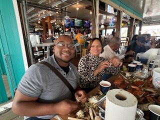 Two people smiling at a seafood market table with crab, drinks, and trays of food