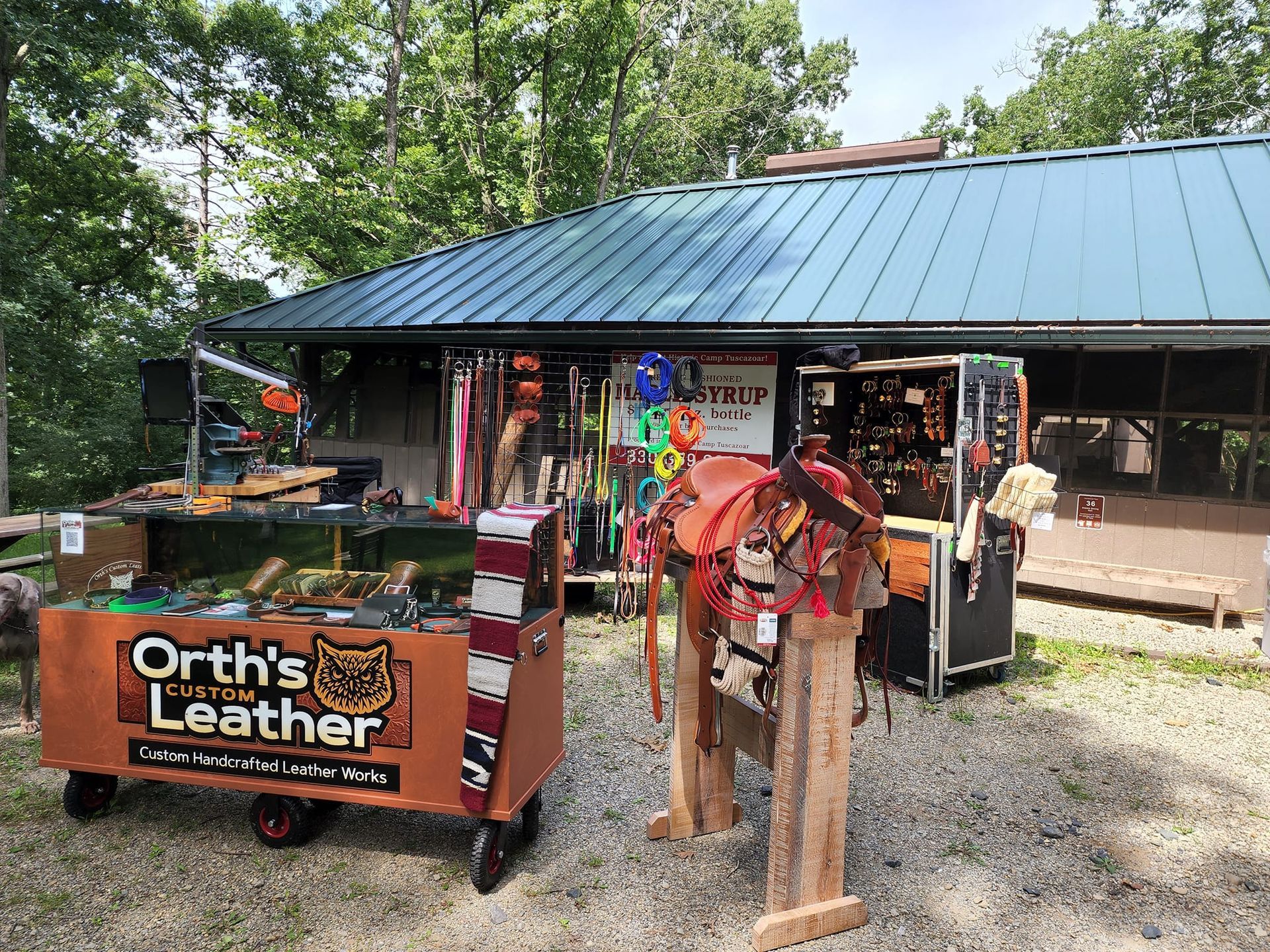 Leather goods booth with an orange “Orth’s Leather” sign and wooden horse saddles on display outdoors