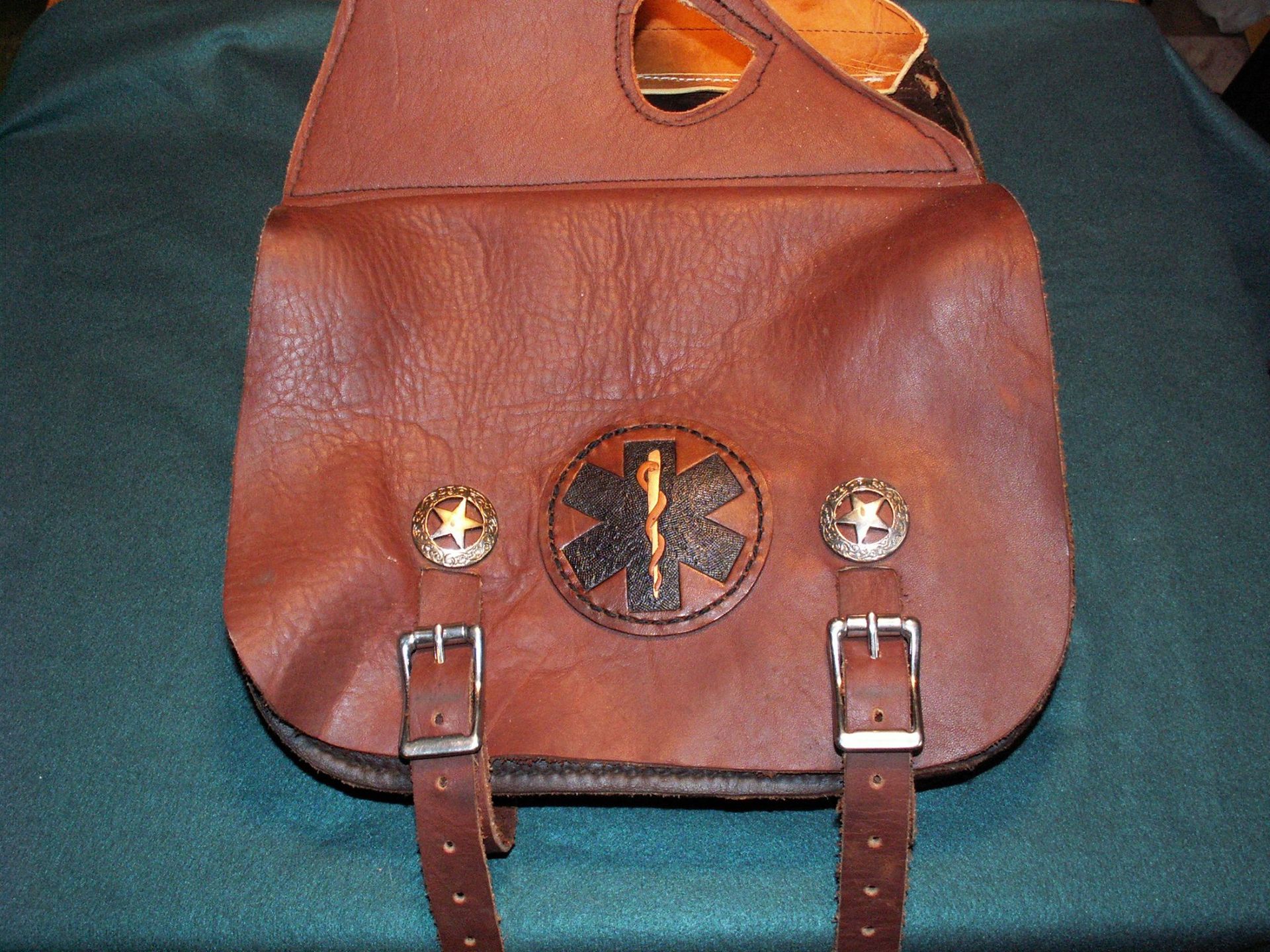 Brown leather shoulder bag with embroidered patch and two buckle straps on a dark fabric background