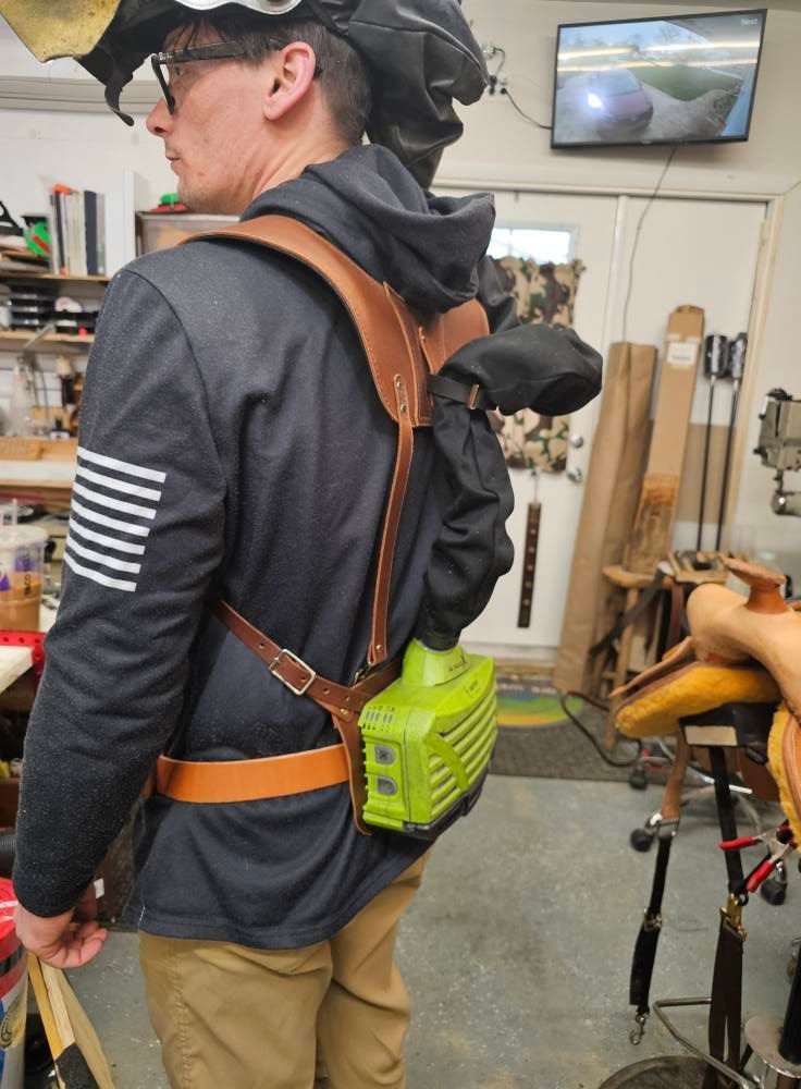 Person in a workshop wearing a dark hoodie, harness, and bright green device on a chair back, with tools nearby