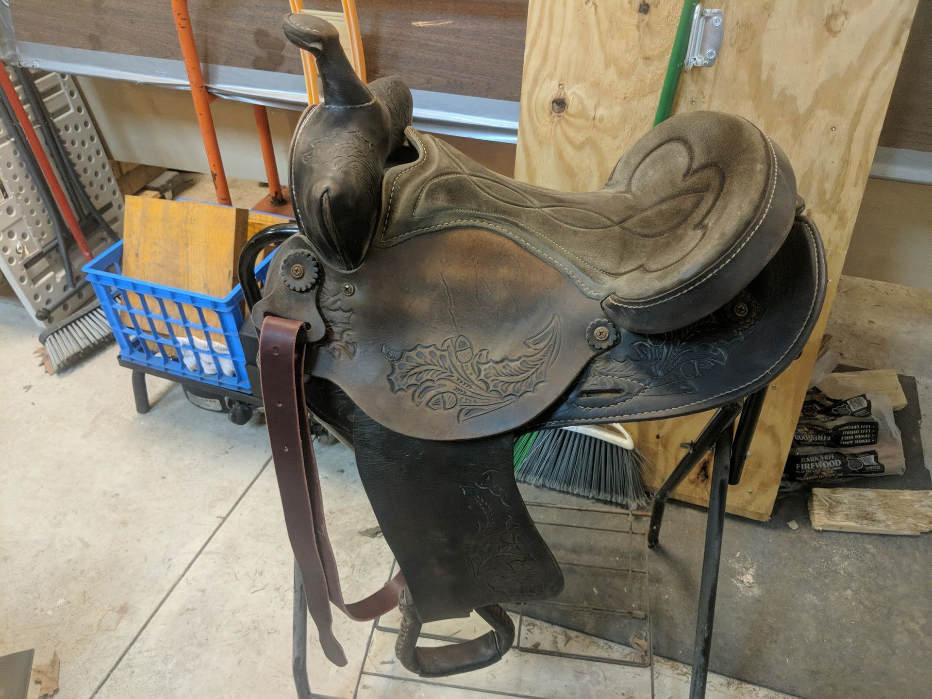 Old brown western saddle on a stand in a workshop, with a blue basket and wooden boards nearby.